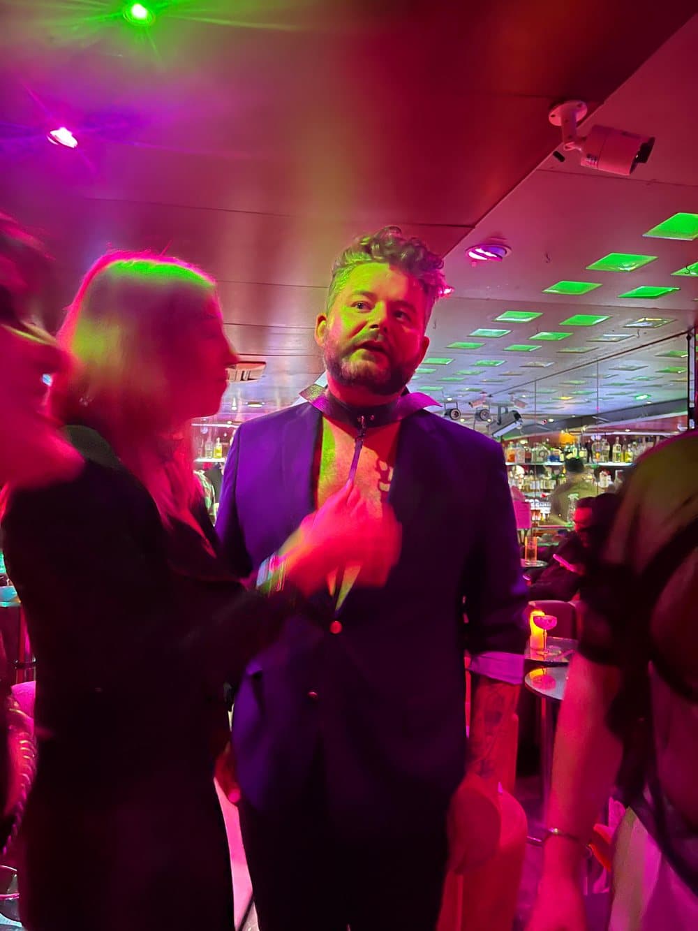 The candidate in a collar and suit at the party, under pink and violet club lights.